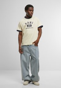 Béžové tričko s námořnickým límcem a grafickým potiskem. Světlé modré volné džíny s viditelným švem. Bílé tenisky, stojící pozice.