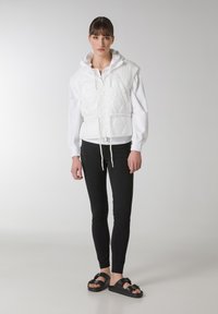Gilet bianco trapuntato con cappuccio, vita con coulisse, indossato sopra una maglietta bianca a maniche lunghe, abbinato a leggings neri e sandali neri.