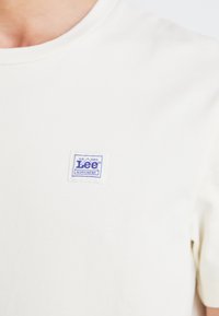 Crèmekleurig t-shirt met korte mouwen, met een vierkant wit label waarop blauwe tekst staat met de woorden "Lee" en "WORKWEAR."