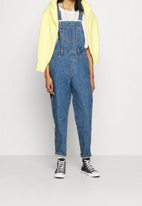 Ljust blå denim-overaller med avslappnad passform, med justerbara axelremmar, ficka fram och uppvikta fållar, i kombination med en gul tröja och svarta sneakers.