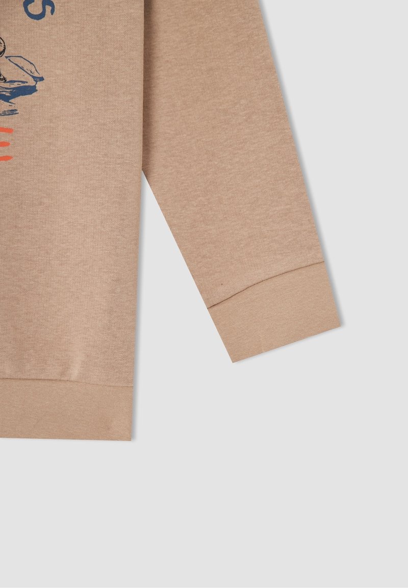 Manchette et ourlet d'un pull beige à manches longues avec un graphique imprimé bleu et rouge partiellement visible sur le tissu, sur fond gris.