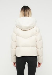 Vrouw met ombre haar, gekleed in een witte gewatteerde jas met capuchon en zwarte jeans, van achteren afgebeeld tegen een eenvoudige achtergrond.