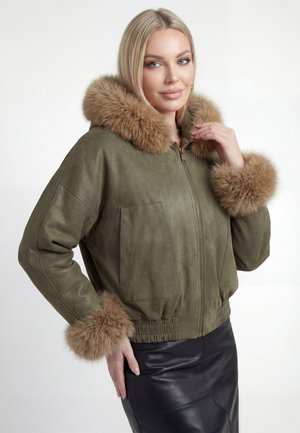 Donna bionda che indossa una giacca verde oliva con rifiniture in pelliccia marrone sul cappuccio e sui polsini, abbinata a una gonna di pelle nera.