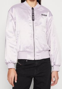 Ljust lila satin bomberjacka med dragkedja, ribbade muddar och nederkant, med en logotypapplikation och sidofickor. Strukturerad, mjuk finish.