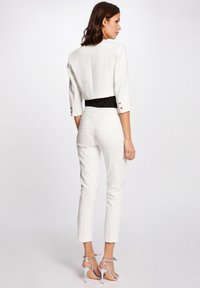 Weißes, tailliertes Blazer mit Dreiviertelärmeln, schwarzen Akzenten und goldenen Knöpfen, kombiniert mit schmalen weißen Hosen und silbernen Riemchenschuhen.