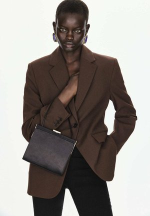 Mujer con blazer marrón y pantalones negros sostiene un bolso negro con una mano en el bolsillo sobre un fondo blanco liso.