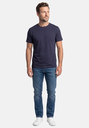 Man standing wearing a navy blue short-sleeve t-shirt, blue jeans, and white sneakers, looking slightly to the right.