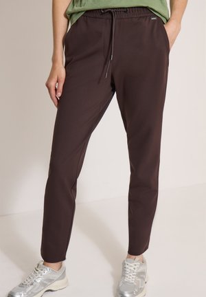 Personne portant un pantalon marron foncé fuselé avec une taille à cordon et des baskets argentées, debout devant un fond clair uni.