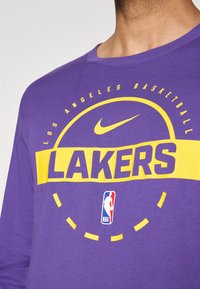Lilla langærmet skjorte med "LAKERS" i fede gule bogstaver, med et Nike-logo ovenfor og et NBA-logo nedenfor, lavet af blødt bomuldsstof.