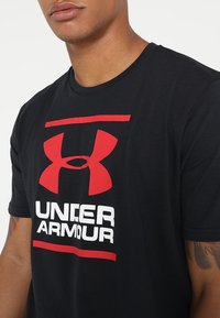 Hombre vistiendo una camiseta negra de Under Armour con un gran logo rojo y texto blanco, mostrando parte de su brazo tatuado.