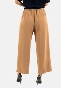Pantalons larges couleur beige en tissu lisse. Dotés d'une ceinture élastique et d'une poche arrière unique. Associés à des talons hauts noirs.