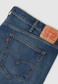 Μπλε τζιν Levi's διπλωμένα, παρουσιάζοντας την πίσω τσέπη με τη ραφή arcuate, το κόκκινο ταμπελάκι Levi's, τη θηλιά της ζώνης και το δερμάτινο ταμπελάκι της μάρκας.