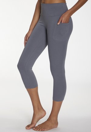 Woman wearing gray high-waisted cropped leggings with side pocket, standing barefoot against plain white background.