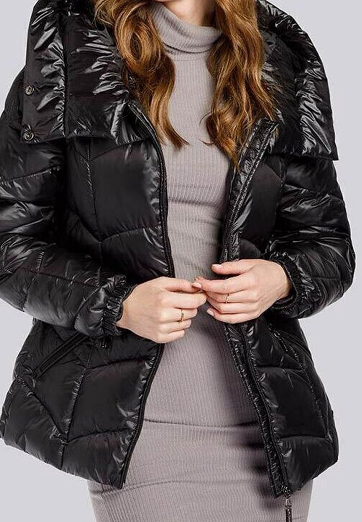 Veste matelassée noire à finition brillante, col surdimensionné, poches zippées et poignets élastiques. Portée sur une robe ajustée grise.