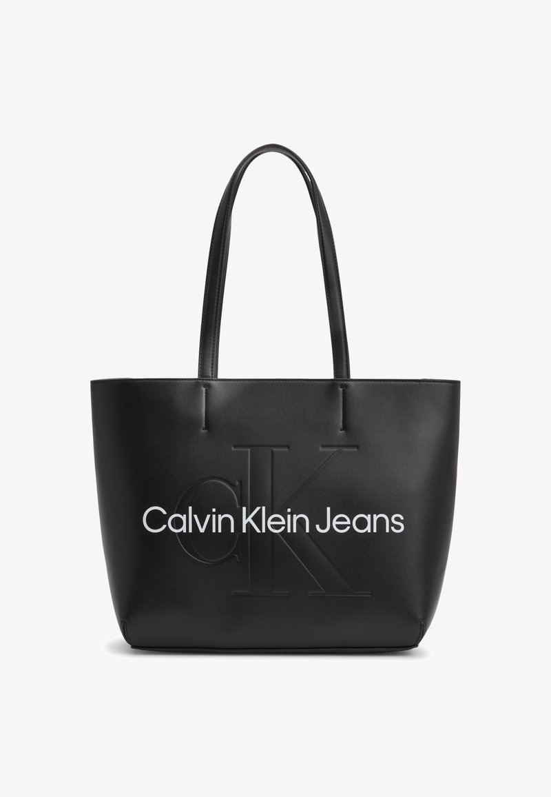 Bolso tote negro hecho de material liso similar al cuero; presenta el logo CK en relieve y texto blanco "Calvin Klein Jeans" en la parte delantera.