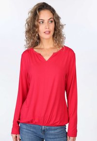 Rotes Langarmoberteil mit V-Ausschnitt und gerafftem Taillenbereich. Glattes Material, lockere Passform und gerippte Akzente. Kombiniert mit blauen Jeans.