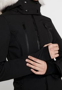 Veste d'hiver noire avec capuche bordée de fourrure, fermeture éclair à l'avant, poches poitrine zippées et poignets avec trou pour le pouce, portée par une personne croisant les bras.