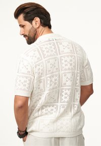 Weißes kurzärmeliges Strickpolo mit einem gemusterten Design, das über ein offenes Gewebe und gerippte Ärmelbündchen sowie Saum verfügt.