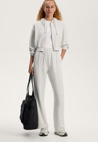 Svetlo šedá skrátená mikina s kapucňou, biele tričko a šedé tepláky s elastickým pásom. Čierna taška, šedé tenisky s akcentmi. Hladké textúry.