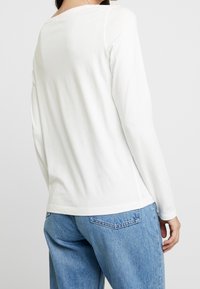 Langärmliges weißes T-Shirt aus weichem Stoff mit einem schlichten Design. Es hat einen runden Ausschnitt und eine lockere Passform, kombiniert mit blauen Jeans.