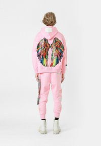 Sudadera rosa con un gran diseño de alas coloridas y de lentejuelas en la parte posterior. Pantalones de chándal rosas a juego con acentos bordados y botas blancas.