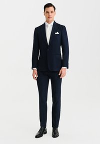 Granatowy garnitur z klapą w kształcie notcha, z pojedynczym zapięciem na guzik i dwoma kieszeniami z przodu. Uzupełniony białą koszulą i krawatem, eleganckie czarne buty.