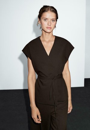 Haut enroulé marron avec des manches courtes, décolleté en V et une ceinture à nouer à la taille. Tissu lisse avec une texture minimale, sur un fond uni.