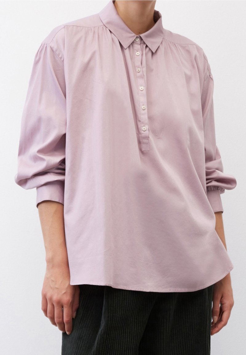 Blusa di un leggero color lavanda con placca bottonata, colletto e dettagli arricciati sulle spalle. Il tessuto appare liscio con una leggera texture lucida.