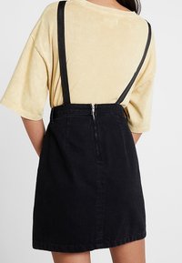 Person som bär en beige kortärmad skjorta och en svart kjol med hängslen, sedd bakifrån mot en neutral bakgrund.