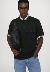 Svart polo-skjorte med hvitstripet krage, utstyrt med en brystlomme. Kombinert med lyseblå denimjeans og en svart crossover-veske.