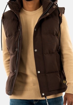 Uomo che indossa un maglione beige a maglia sotto un gilet imbottito marrone scuro con bottoni a pressione argentati e zip, abbinato a jeans blu.