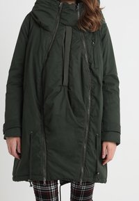 Žena v tmavozeleném zatepleném zimním kabátu s několika zipy, velkou kapucí a kostkovanými kalhotami na jednobarevném pozadí.