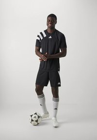 Uniforme de fútbol atlético negro con detalles blancos, incluyendo rayas en las mangas y los pantalones cortos. Presenta un logo de Adidas. Jugador posando con un balón de fútbol.