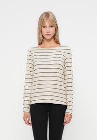 Beige hosszú ujjú felső vékony fekete csíkokkal, kerek nyakkivágással, laza fazonnal, fekete bordás leggings-szel párosítva. Puha textúra, laza dizájn.