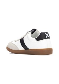 Zapatilla blanca con rayas negras y una capucha negra en el talón, con una suela de goma texturizada y una puntera de ante. Cordoneras en un estilo clásico.