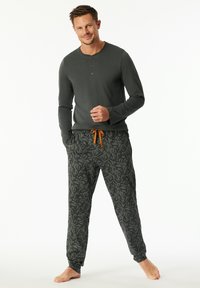 Man standing barefoot wearing dark green long-sleeve henley shirt and patterned drawstring jogger pants with orange tie.