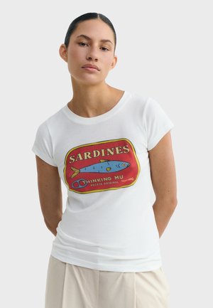 Femme aux cheveux foncés tirés en arrière, portant un t-shirt blanc avec un motif de boîte de sardines rouge, jaune et bleu, et un pantalon beige.