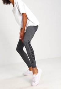 Grå leggings med en glansig yta som har en svart "Superdry"-logotyp på vänster ben, kombinerat med en lösvit topp och vita sneakers.