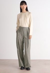 Kremna bluzica s naboranim izrezom, kombinirana s sivo prugastimi širokimi hlačami. Črne čevlje dopolnjujejo outfit. Minimalističen dizajn, mehak material.