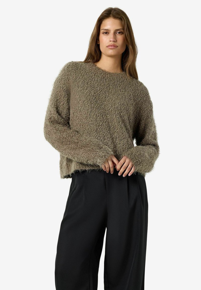 Femme aux cheveux longs porte un pull gris duveteux et un pantalon noir ample, debout devant un fond uni, les mains délicatement jointes.