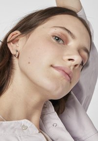 Pendientes de plata de ley con un diseño texturizado. El modelo lleva una camisa de color púrpura claro con botones a presión de metal.