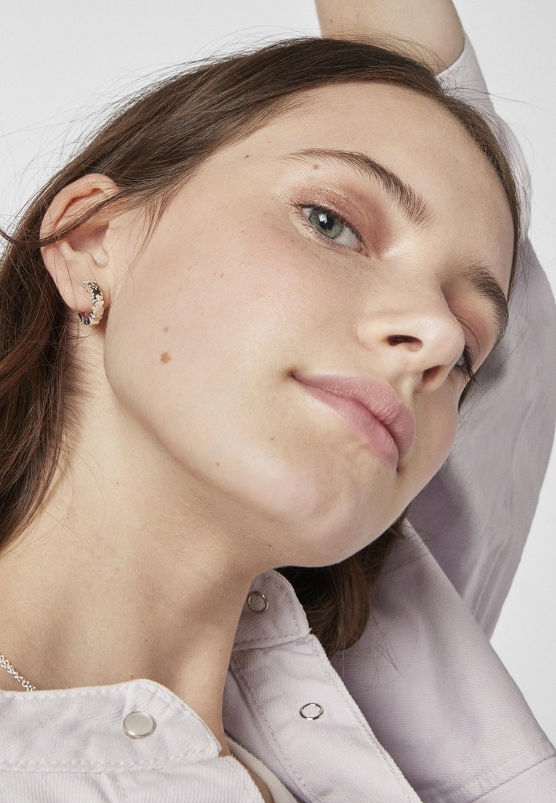 Pendientes de plata de ley con un diseño texturizado. El modelo lleva una camisa de color púrpura claro con botones a presión de metal.