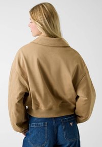 Tan korte jas met brede kraag, zachte textuur en geribbelde zoom, gecombineerd met blauwe high-waisted spijkerbroek met achterzakken.