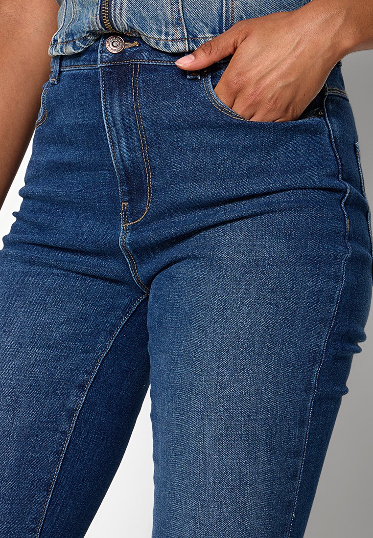 Main dans la poche d'un jean en denim bleu taille haute avec des coutures visibles et un bouton en cuivre, porté avec une veste en denim assortie.