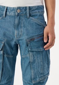 Pantalones cargo de mezclilla en azul claro con múltiples bolsillos, un bolsillo lateral con cremallera y un cierre de botón en la cintura. Tejido texturizado con áreas desgastadas.