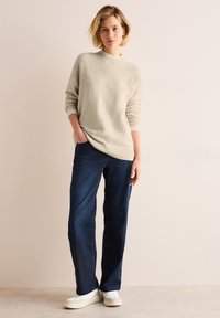 Beige gestrickter Pullover mit Ripptextur, kombiniert mit dunkelblauen geraden Jeans und weißen Sneakers, vor einem neutralen Hintergrund.
