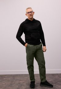 Sort hættetrøje med en forlommes, olivengrønne bukser og sorte sneakers. Looket har et afslappet design med bløde teksturer.