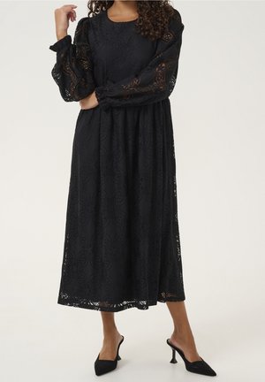 Kobieta w długiej czarnej koronkowej sukni z bufkami z przezroczystego tiulu i czarnych, szpiczastych mule na obcasie na jednolitym tle.