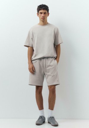 Mladić stoji naslonjen na bijeli zid noseći svijetlosivu oversized majicu, odgovarajuće kratke hlače, bijele čarape i sive tenisice, s rukama u džepovima.