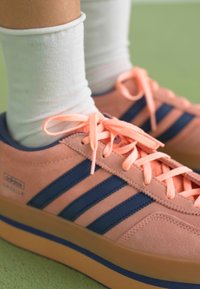 Svetlo roza semšaste tenisice s tremi temno modrimi črtami, roza vezalkami in debelim gumijastim podplatom, oblečene z belimi nogavicami do gležnjev.